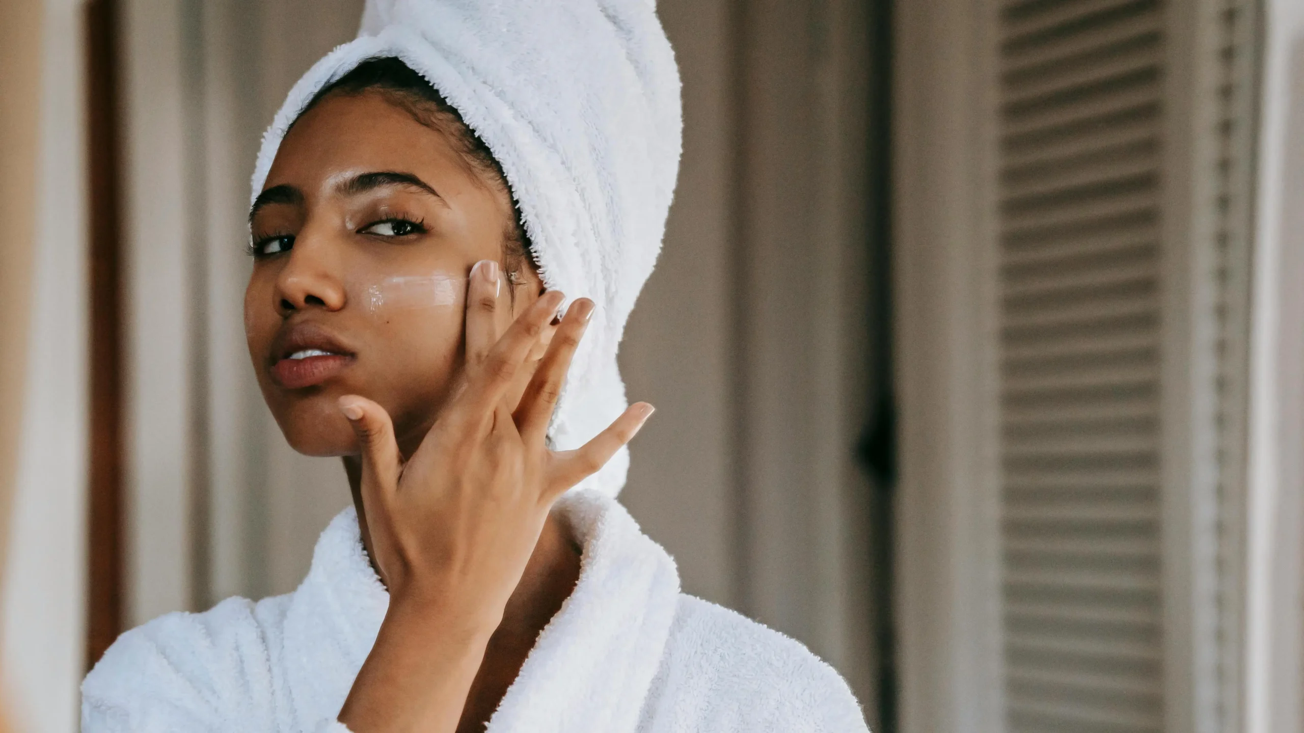 Young woman in robe with towel on head applying chub rub cream on face in front of mirror