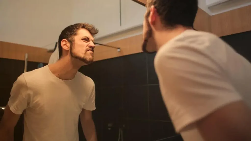 Young man in casual shirt frowning in bathroom mirror, concerned about odor despite showering