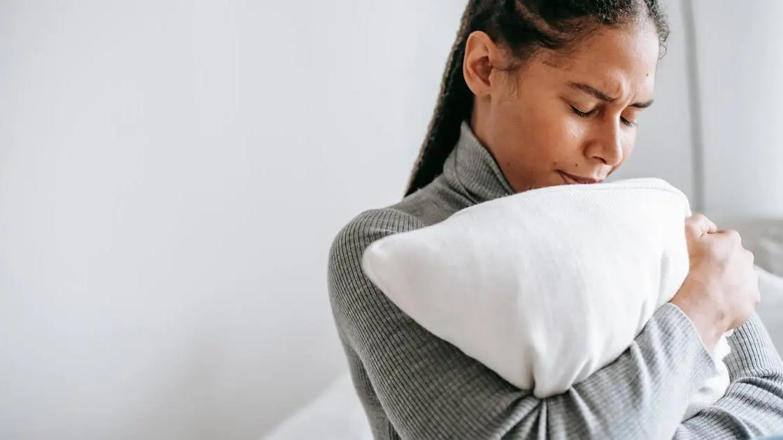 Young woman with closed eyes, looking distressed, sitting on bed and hugging pillow, reflecting Psychological Impact of Night Sweats