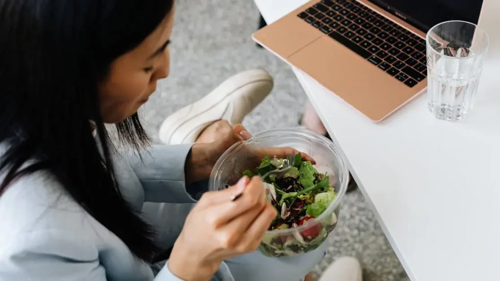 Woman enjoying fresh salad at desk with laptop, illustrating GLP-1 Weight Loss Programs and healthy eating.